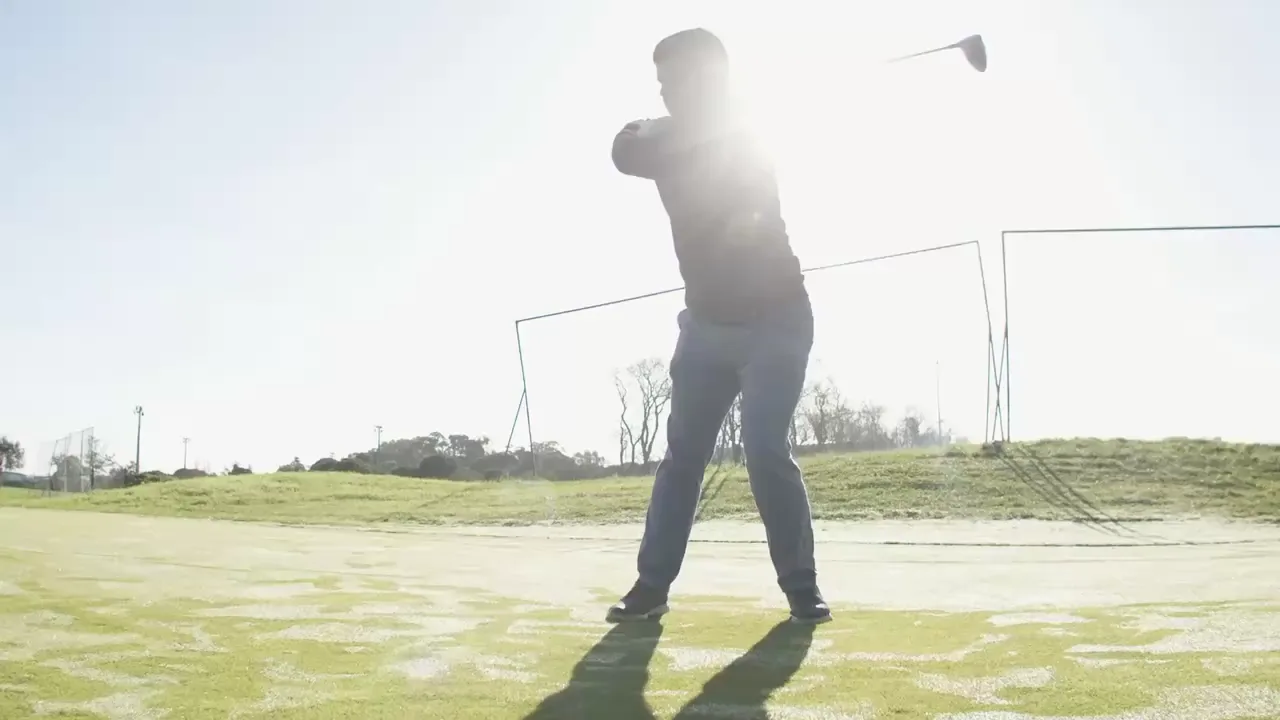 A man driving a golf ball on a sunlit practice range